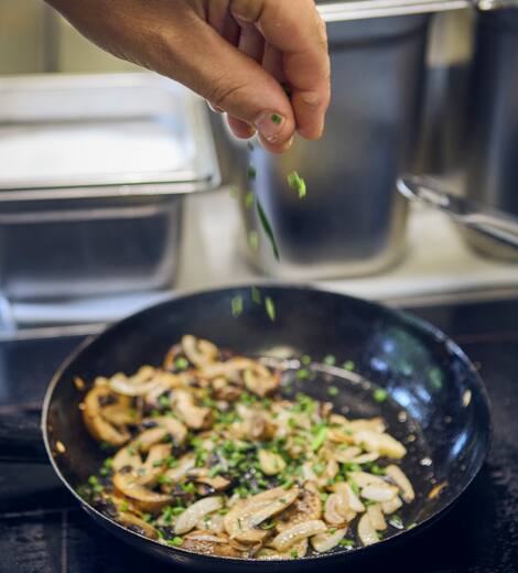 Koch streut frische Petersilie über gebratene Champignons in der Pfanne, direkt in der Hotelküche